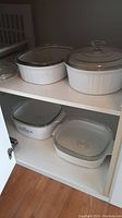 Four Corning baking dishes in a shelf cupboard, two on each shelf, two round white ribbed dishes with lids on upper shelf and two square dishes, one with blue floral pattern on lower shelf.