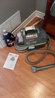 Full view of Bissell SpotBot carpet cleaner unit on hardwood floor with attached hose, cleaning solution bottle, and instruction manual next to it.