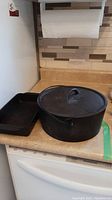 Cast iron pot with lid and bail handle alongside rectangular cast iron baking pan on kitchen counter.