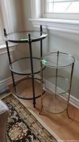 Two round three-tiered display tables, one with black metal frame and one with brass-colored metal bamboo-style frame, both with clear glass shelves.