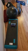 Photo showing black and teal exercise step platform, purple and blue dumbbells, three rolled yoga mats, and large black balance ball on wooden floor.