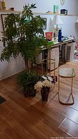 Large faux tree, two faux flower arrangements in pots, round gold-colored side table with missing glass top, and rectangular white-top side table all positioned on living room floor.