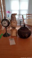 Full view of all three items on tabletop with window background: marble bird statue, round clock on brass stand, ebony carved wooden bowl.