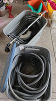 Two large gray plastic bins, each containing a coiled gray garden hose; one bin includes a water wand attachment on top of the hose.