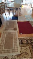Overview of various rugs and mats laid out on wooden floor including beige rugs, burgundy mat, blue mat.