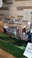 Side view of shiny silver-tone Airstream camper-shaped teapot with painted colorful windows and door, sitting on original green box.