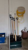 Four shovels with various handle types and sizes, standing side by side with a yellow extension cord hanging on the wall above a wall hook.