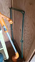 Orange and black Worx yard trimmer leaning against a wooden wall, green metal guard stand beside it.