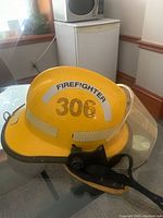 Yellow firefighter helmet showing front view with 'FIREFIGHTER 306' label and attached face shield and chin strap, placed on a table indoors.