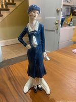 Full view of the vintage Goebel flapper girl figurine dressed in blue, standing on a white base on a wooden table indoors.