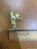 Gold-colored Mickey Mouse trinket figurine placed next to a ruler showing size approximately 2 inches tall, with visible damage on the right ear.