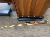 Two disassembled pool cues laid out with both a soft vinyl case and a hard case beneath a wood-finish desk