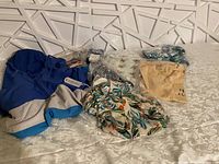 Overview of multiple new clothing items from Beachly including a blue rain hoodie, tropical printed shirt, beige t-shirt, slippers, and other packaged items spread on a bed.