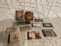 The lot shows a collection of 12 CDs and DVDs arranged on a patterned white tablecloth. Some cases are new and unopened, others show wear.