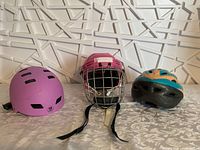 Front view showing all three helmets: a purple bike helmet, a pink hockey helmet with face guard, and a teal and orange bike helmet.