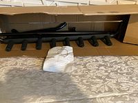 Unassembled black metal guitar stand parts including tubular frame and padded holders inside open cardboard box, with installation guide placed on front covering parts.