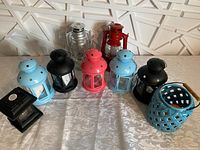 Photo of various decorative lanterns arranged on a patterned cloth against a white tiled backdrop. Includes metal and ceramic lanterns in blue, black, red, pink, silver, and teal colors.