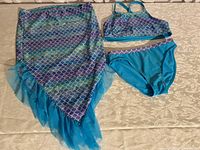 Overall shot of mermaid swimsuit outfit including tail skirt, swim top and bikini bottoms laid out on cream patterned surface.