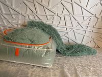 Green shaggy comforter and pillow sham partially visible in transparent plastic storage bag with orange zipper and handle, fibers having a soft and plush shaggy texture.