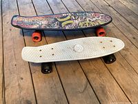 Top view of two skateboards stacked; top skateboard has colorful graffiti style deck and orange wheels, bottom skateboard is white plastic cruiser style with textured grip and black wheels.
