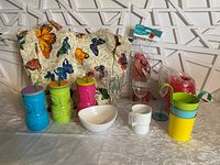Various summer-themed items on table including colorful drink tumblers with lids and straws, ceramic bowl and cup, wine glasses, red heart candle, butterfly cushion, and packaged decorative item.