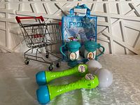 Metal toy shopping cart with red handle, two blue Frozen-themed cups, two green bubble wands, and blue Frozen tote bag arranged on tabletop against wall