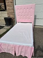 Full view of twin bed showing soft pink upholstered headboard, white mattress cover and matching pink bed skirt, outdoors against garage door.