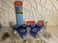 Winter-themed decor items including six blue metal snowflake tins, packaged snow blanket fabric, silver mini ornaments, frosted votive candle holders, and snowflake tealight set on silver tablecloth.