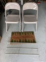 Two beige metal folding chairs with minor dents, a Christmas-themed coir doormat with green tree silhouettes and "Happy Holidays" text, and a white wire shelf.