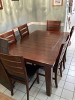 Photo of rectangular wooden dining table with six matching chairs surrounding it, showing plank-style top and dark wood finish.