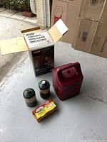 Photo shows Coleman camping lantern box, two Coleman propane canisters with white caps, red carrying case, and a yellow box of Sterno fuel cans on concrete surface near garage door.