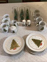 Photo showing entire 26-piece Christmas dish set arranged on white quilted surface includes coffee cups, creamer, sugar container, salt and pepper shakers, clear glass water glasses with Christmas trees inside, and dessert plates with Christmas tree and gold trim.