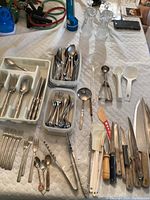 Top down view of bamboo pattern flatware set, various knives, and kitchen utensils on white quilted surface including forks, spoons, knives, cocktail forks, tongs, melon ball scoop, ladles, and small spoons.