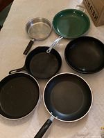 Photo showing five frying pans in a group: one cast iron skillet, one green non-stick pan, one stainless steel pan, and two black non-stick pans. Items show scratches and wear.