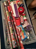 Top-down view of plastic container with assorted Christmas wrapping paper rolls, gift ribbons, and bags.
