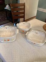 Three Corning Ware casserole dishes on a table with white textured tablecloth. All have the floral Wildflower pattern featuring orange and yellow flowers. Each has a clear glass lid with a round knob. The pieces vary in size: one large oval or square, one medium square, and one rectangular smaller casserole dish.