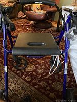 Front view of Drive sit down walker showing blue frame, black seat with carrying hole, and wheels on carpeted floor.