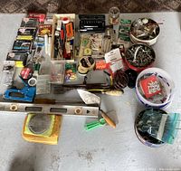 Overview shot showing all tools and small hardware items arranged on floor including level, crowbar, screwdrivers, socket set, and multiple small containers of screws, nails, and anchors.
