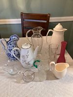View of the 11 assorted pitchers, vases, creamers, and teapot on table including ceramic, porcelain, and glass pieces of varying sizes and colors.