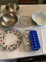 Displays all items in the lot on a white textured cloth on table: two aluminum bowls, white ceramic bowl, glass candy dish, glass serving platter with floral decoration, blue ice cube tray, metal hot plate holder, green Pyrex bowl