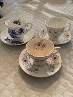 Image showing three different floral pattern teacups each with matching saucers laid on quilted fabric. One cream with blue and pink flowers and gold trim (Royal Kent), one blue floral pattern on white (Avon), and one multi small floral prints on white (Crown)