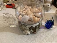 Photo of glass bowl filled with shells on decorative base, flanked by two glass fish figurines, on a white tablecloth.