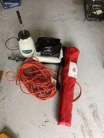 Photo shows Scotts branded pump sprayer, black folded plastic tarps, orange extension cords coiled, and a red portable quad chair in carrying case on concrete floor.