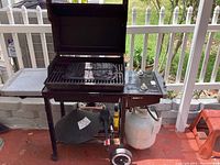 Weber gas grill with open lid showing cooking area and cast iron grill pan. Side control panel with knobs and propane tank below mounted on wheeled frame. Grill tools hanging on lower shelf.