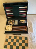 Opened black carrying case showing backgammon board on lid, chess pieces, cribbage boards, dominoes and playing cards inside compartments.