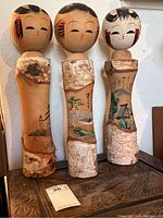 Front view of three tall birch log Kokeshi dolls with painted faces and scenic decorations on birch log bodies