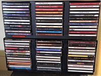 Photo of rock and jazz CD collection in plastic cases stacked vertically in a black storage case. Artist names and album titles visible on spines.