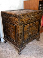 Full view of the carved wood chest showing left and front panels, decorative floral carvings, brass latch, and short carved legs.