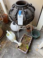 Large clay pot with metal chain decoration on white metal stand, visible ground arrangement showing multiple pots and watering cans