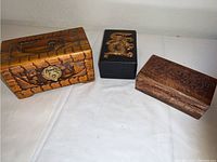 Three hand carved wooden boxes displayed on white fabric: largest box with scenic carving and metal clasp on the left, black box with gold dragon in center, smaller floral carved wood box on right.